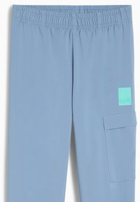 Pantalones deportivos de color azul claro con cintura elástica, bolsillo lateral y un pequeño parche de logo en verde menta. Textura de tela suave y ligera.
