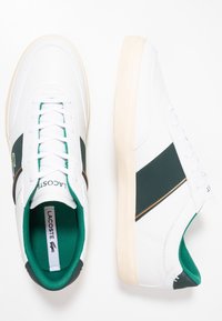 Bílé tenisky se zelenými detaily, s hladkým koženým svrškem, plochými tkaničkami, polstrovaným límcem a logem značky na boku.
