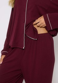 Completo loungewear bordeaux composto da due pezzi, con una camicia a maniche lunghe con bottoni e pantaloni con vita elastica, caratterizzato da piping bianco e texture a costina.