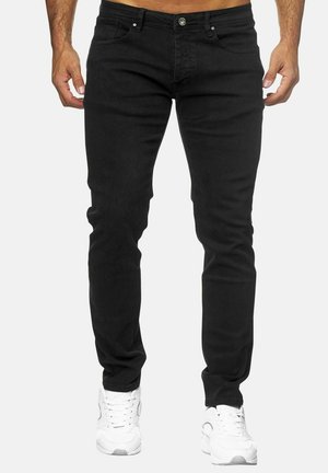 Hombre vestido con vaqueros negros ajustados y zapatillas blancas, de pie frente a un fondo claro y liso, con las manos relajadas a los costados.