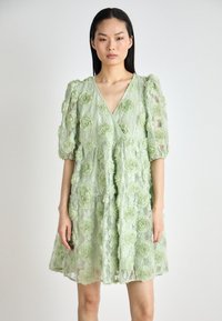 Robe en dentelle verte avec des appliqués floraux, décolleté en V, manches bouffantes et design superposé. Tissu texturé avec une finition transparente.