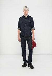 Mørkeblå denimskjorte med to brystlommer, rullet ermer og matchende bukser. Svarte snøresko og en rød caps i hånden.