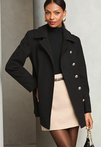 Cappotto di lana nero con bottoni argentati, design con colletto, abbinato a un dolcevita nero a coste e una gonna beige; collant neri e borsa.