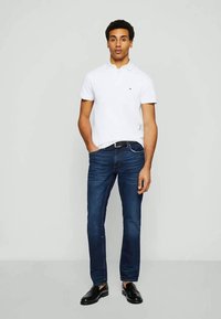 Man som står med vit kortärmad polo, mörkblå jeans, svart bälte och svarta loafers mot en enkel ljusgrå bakgrund.