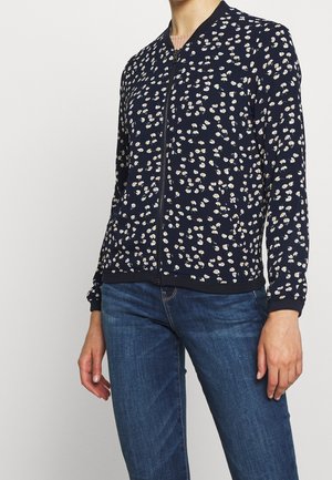 Femme portant une veste zippée bleu marine avec un petit motif floral blanc et beige et un jean bleu, debout devant un fond uni.