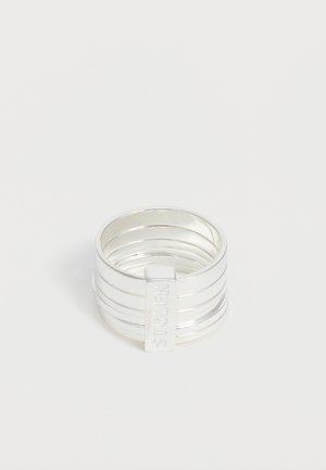 Ασημένιο πλατύ δαχτυλίδι με οριζόντιες αυλακώσεις και κάθετη ορθογώνια πλάκα με ανάγλυφο το "GUESS" σε μονόχρωμο φόντο.