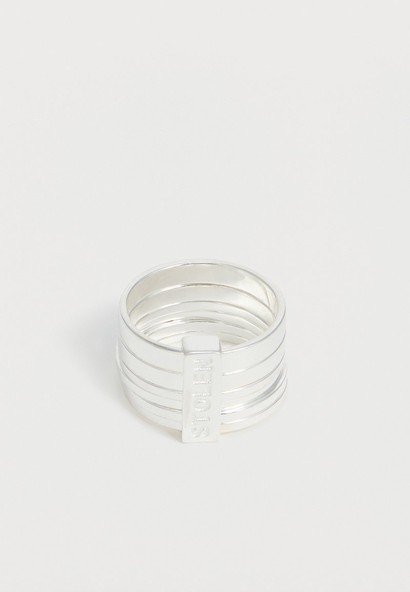 Ασημένιο πλατύ δαχτυλίδι με οριζόντιες αυλακώσεις και κάθετη ορθογώνια πλάκα με ανάγλυφο το "GUESS" σε μονόχρωμο φόντο.