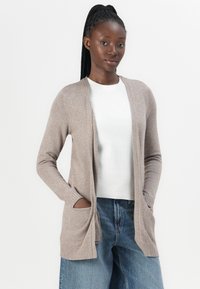 Beige öppet fram cardigan tillverkad av mjukt material, med långa ärmar, ribbade detaljer och två framfickor, bärs över en vit topp.