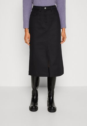 Vrouw draagt een zwarte midirok met een split aan de voorkant, gecombineerd met zwarte kniehoge laarzen en een lila trui met lange mouwen, staand tegen een effen achtergrond.