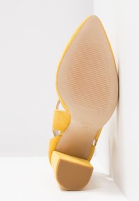 Chaussure à talons hauts en daim jaune avec un bout pointu, dotée d'une sangle transparente, d'une semelle texturée et d'un design de talon plat.