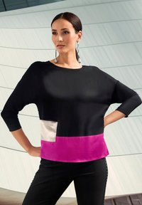 Frau mit dunklen Haaren in einem schwarzen Oberteil mit beigem Taschen und pinkem Saum, die mit auf die Hüften gestützten Händen vor einer modernen Wand steht.