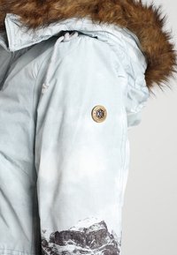 Svetle modrý parka kabát s kožíškovou kapucí, s potiskem hor na rukávu a kulatou nášivkou na paži.