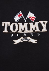 Černá látka s vyšitým logem "TOMMY JEANS", obsahující dvě vlajky a banner s nápisem "EST 1985" v béžové a bílé barvě.