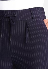 Hose in marineblau mit kariertem Muster, Gummizugbund, Gürtelschlaufen, seitlichen Falten und einer Hand in der rechten Tasche.