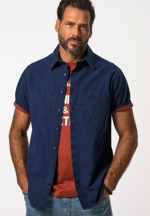 Short-sleeve, button-up shirt in dark navy denim with two chest pockets, layered over a rust-colored graphic T-shirt, worn with light jeans.