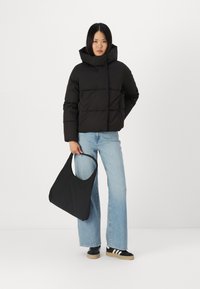 Zwarte pufferjas met hoge kraag en rechthoekig doorgestikt ontwerp, gecombineerd met lichtblauwe flared jeans en een zwarte tote bag.