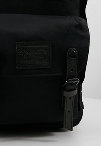 Sac à dos en tissu noir avec poche avant à rabat, écusson logo rectangulaire cousu et sangle noire réglable avec boucle et fermeture par bouton-pression.