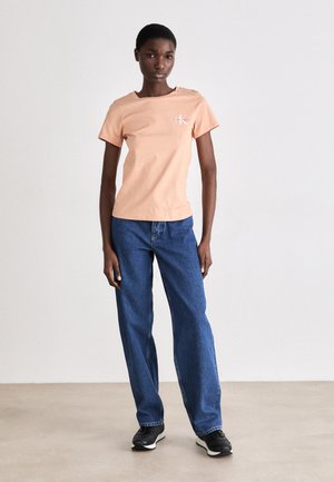 T-shirt léger pêche à manches courtes avec un petit logo, associé à un jean bleu ample et des chaussures de sport noires sur un fond neutre.