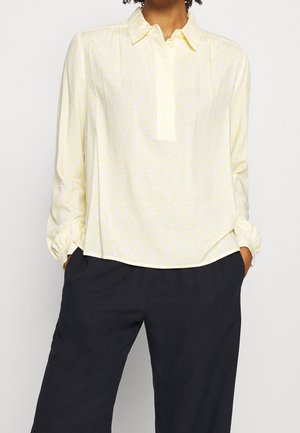 Kobieta w jasnożółtej kwiecistej bluzce z długim rękawem i kołnierzykiem oraz czarnych szerokich spodniach, z rękami w kieszeniach, stojąca na jednolitym tle.