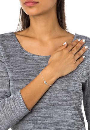 Zilveren kettingarmband met een kleine vleugelbedel, gedragen om een pols; model heeft een grijs textuur langemouwen top en neutrale nagellak.