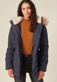 Chaqueta acolchada azul marino con capucha forrada de pelo sintético, cierre de cremallera y botones a presión. Presenta puños acanalados y bolsillos laterales. Se lleva sobre una blusa marrón.