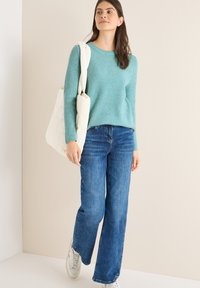 Helles türkisfarbenes geripptes Sweatshirt, blaue weit geschnittene Jeans und weiße gestepfte Tasche. Model trägt graue Sneaker und steht vor einem neutralen Hintergrund.