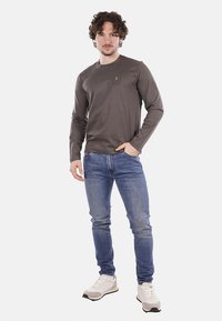 Szara koszula z długim rękawem z kieszonką na piersi i logo, w połączeniu z niebieskimi dżinsami i białymi trampkami. Koszula ma gładką fakturę materiału.