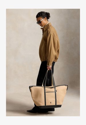 Femme portant une veste marron et un pantalon noir, tenant un grand cabas beige avec des anses et des bordures noires, debout sur un fond uni.