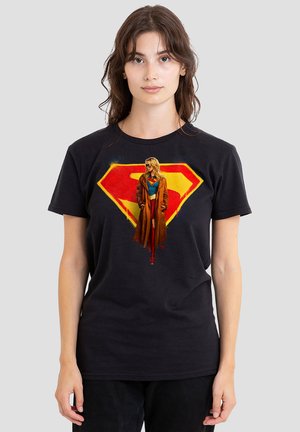 Frau trägt schwarzes T-Shirt mit rotem und gelbem Superman-Logo und einer Supergirl-Figur in einem braunen Mantel, die in der Mitte aufgedruckt ist.