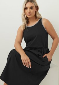 Svart ärmlös klänning med elastisk midja, prydd med vit dekorativ kant längs halsringningen och sidofickor. Tillverkad av ett mjukt tyg.