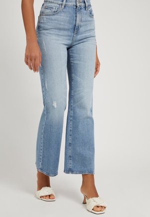 Vrouw draagt lichtblauwe, hoge taille, rechtgesneden jeans met lichte slijtage en witte sandalen met hak en open teen, tegen een effen achtergrond.
