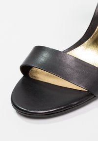 Sandale noire à bout ouvert avec une large bride en cuir sur les orteils et une semelle intérieure de couleur dorée visible à l'intérieur de la chaussure.