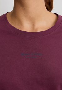 Primer plano de una persona que lleva una camiseta de cuello redondo morada con "Marc O'Polo EST. EN ESTOCOLMO" impreso en letras oscuras en el pecho.