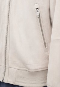 Veste beige clair avec une texture douce en daim. Dispose d'une poche zippée, d'un ourlet côtelé et de détails de couture. Design minimaliste.