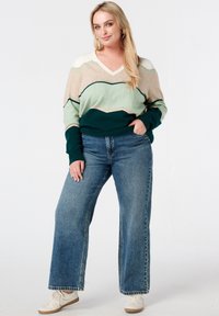 Pull en tricot à col en V, avec des rayures en zigzag beige, vert et vert foncé ; associé à un jean large taille haute en denim et des baskets blanches.