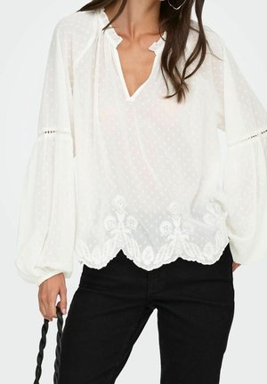 Femme portant un chemisier blanc à manches longues avec une broderie en dentelle et un pantalon noir, tenant un sac à main à poignée torsadée noire.