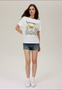 Jovem mulher com cabelo castanho longo, vestindo uma t-shirt branca com estampado de borboletas e flores, calções de ganga e sapatilhas brancas, de pé contra um fundo simples.