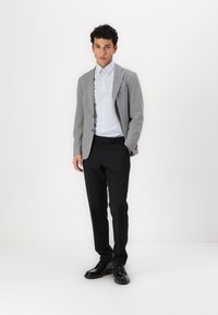 Blazer grigio, camicia a righe azzurro chiaro, pantaloni neri e scarpe nere. Abbigliamento formale su uno sfondo bianco uniforme.
