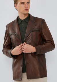 Blazer de cuero marrón, con cuello con muescas, cierre frontal de botón y bolsillos en el pecho con cremallera. Textura suave y diseño ajustado.