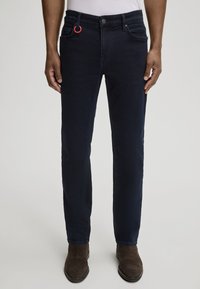 Granatowe dżinsy o slim fit, z czerwonym akcentem w postaci pierścienia na szlufce paska oraz subtelnie wystrzępioną teksturą.