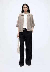 Femme portant un cardigan beige à manches courtes boutonné, haut blanc, pantalon noir à jambes larges, tenant un sac à main beige clair, debout devant un fond uni.