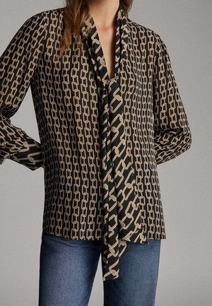 Femme portant un chemisier noir avec un motif beige de maillons de chaîne et une écharpe assortie, associée à un jean bleu, debout devant un fond gris.