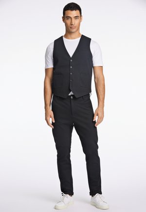 Chaleco negro sobre una camiseta blanca, abrochado con dos bolsillos frontales. Combinado con pantalones negros ajustados y zapatillas blancas. Fondo liso.