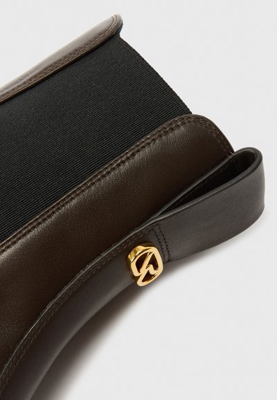 Καφέ δερμάτινη τσάντα με μαύρη ελαστική πάνελ. Περιλαμβάνει ένα χρυσό λογότυπο από υλικό και έχει απαλό υφές, σχεδιασμένη με στρογγυλεμένες γωνίες.