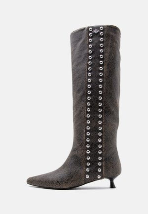Bota alta de cuero negro con tacón bajo, punta puntiaguda y filas verticales de tachuelas plateadas a lo largo de una tira de ante en el lateral.