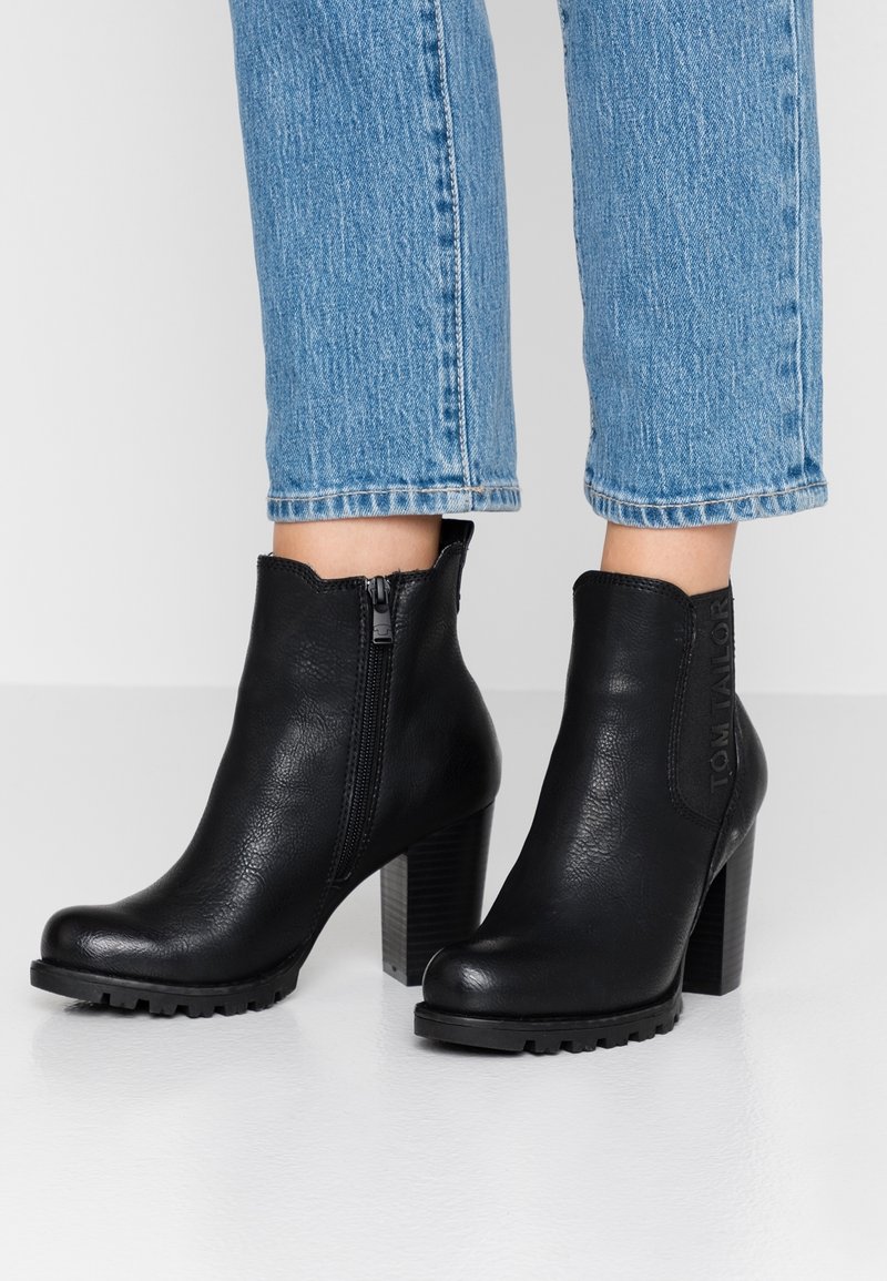 Bottes cavalières en cuir noir avec talons blocs et fermetures éclair latérales, portées avec un jean bleu court sur un fond blanc.