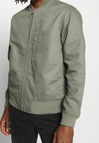 Zielona oliwkowa bomberka wykonana z gładkiego materiału, z zamkiem błyskawicznym z przodu, ściągaczami na mankietach i kieszenią na piersi. Prosty, elegancki design.