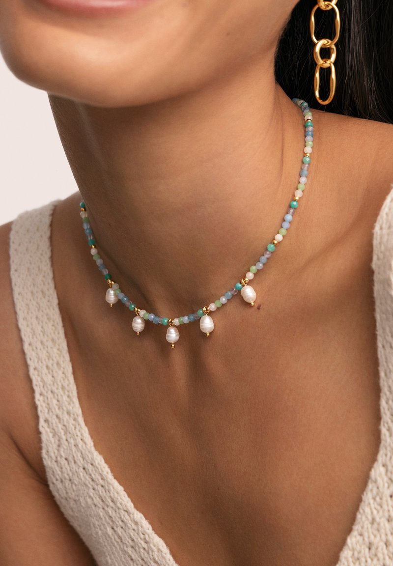 Bewerkte choker-ketting met iriserende parels, meerkleurige glazen kralen in blauw en groen, en gouden accenten, rustend op de huid.