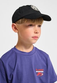 Svart baseballkeps med cirkulär logotyp-patch, i kombination med en lila t-shirt med röda grafikdetaljer. Fokus på tygtextur och färger.