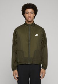 Lekka kurtka w kolorze oliwkowej zieleni z wysokim kołnierzem, zapięciem na suwak, bocznymi kieszeniami i białym logo na piersi. Gładka, nylonowa faktura.